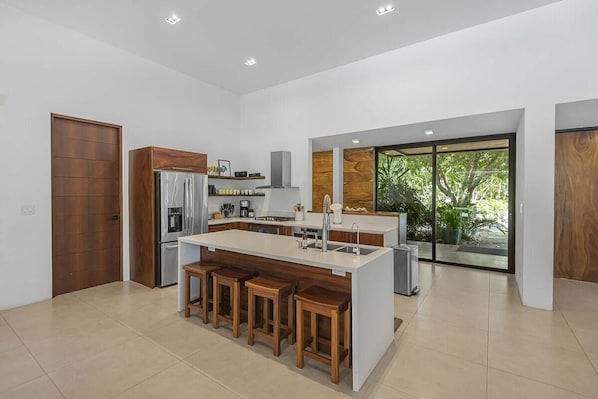 A modern kitchen with a central island, bar stools, and a large window providing a view of greenery.