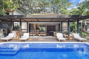 Un espace aménagé au bord de la piscine, avec des chaises longues, un coin salon abrité et une vue sur l’intérieur.