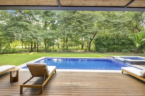 A wooden deck with two lounge chairs and a small table, overlooking a swimming pool and a garden with trees.