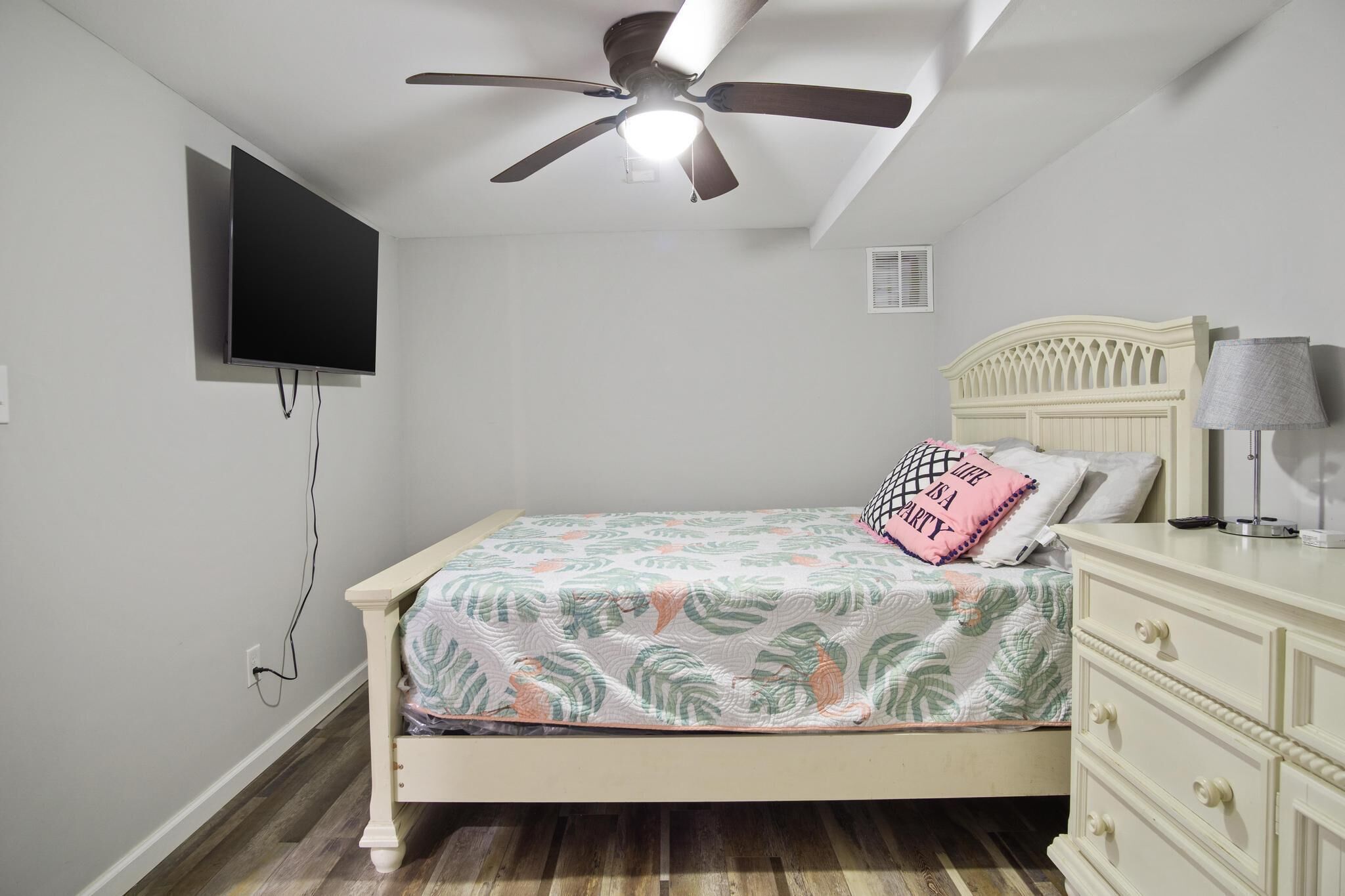 A bedroom with a bed, a ceiling fan, a TV mounted on the wall, a dresser, and a lamp.