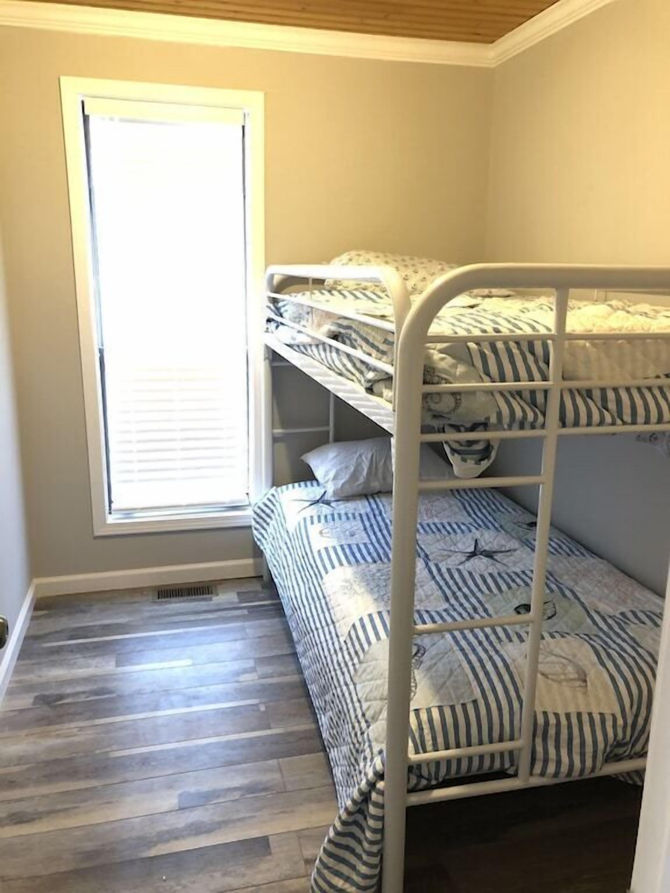 Une chambre avec un lit superposé, dotée de draps à rayures, d’une fenêtre et d’un plancher en bois.