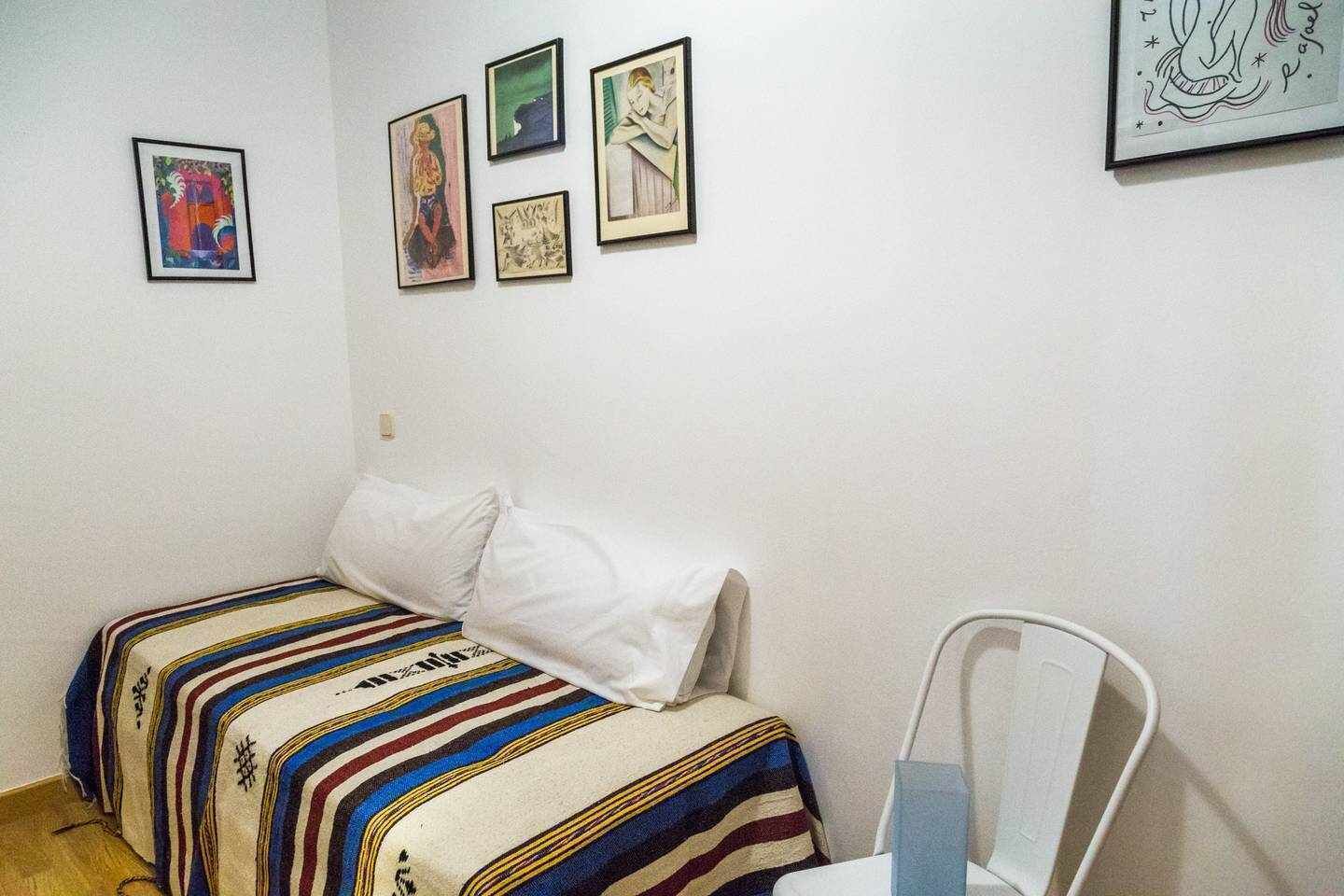 Una habitación pequeña y sencilla con una cama individual, una silla y cuatro obras de arte enmarcadas en la pared.