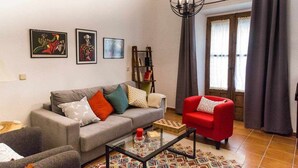 Una sala de estar con un sofá gris, un sillón rojo y una mesa de centro. Hay tres obras de arte enmarcadas en la pared, una estantería y una araña.