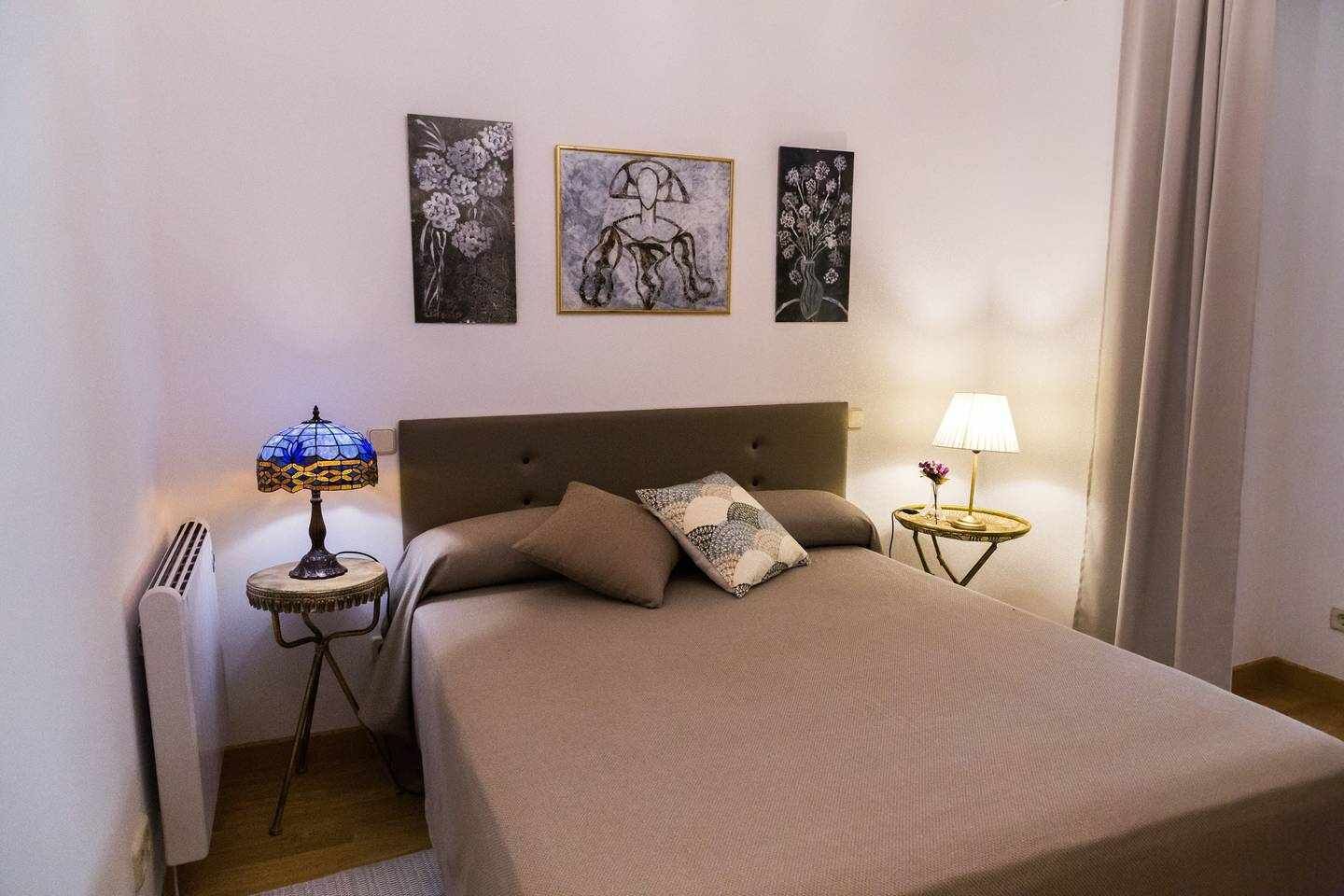 Une chambre à coucher avec un lit, deux tables de chevet avec des lampes, un radiateur et trois œuvres d’art encadrées accrochées au mur.