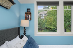 A bedroom with a bed, a bedside table with a lamp, a bookshelf, and a window with a view of a house and trees. - Muskoka Night Dreams: Enjoy 3 King Beds! (Huntsville)