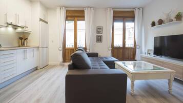 Ein modernes Wohnzimmer mit einer Couch, einem Fernseher und einer Küchenzeile inklusive Schränke und Kühlschrank.
