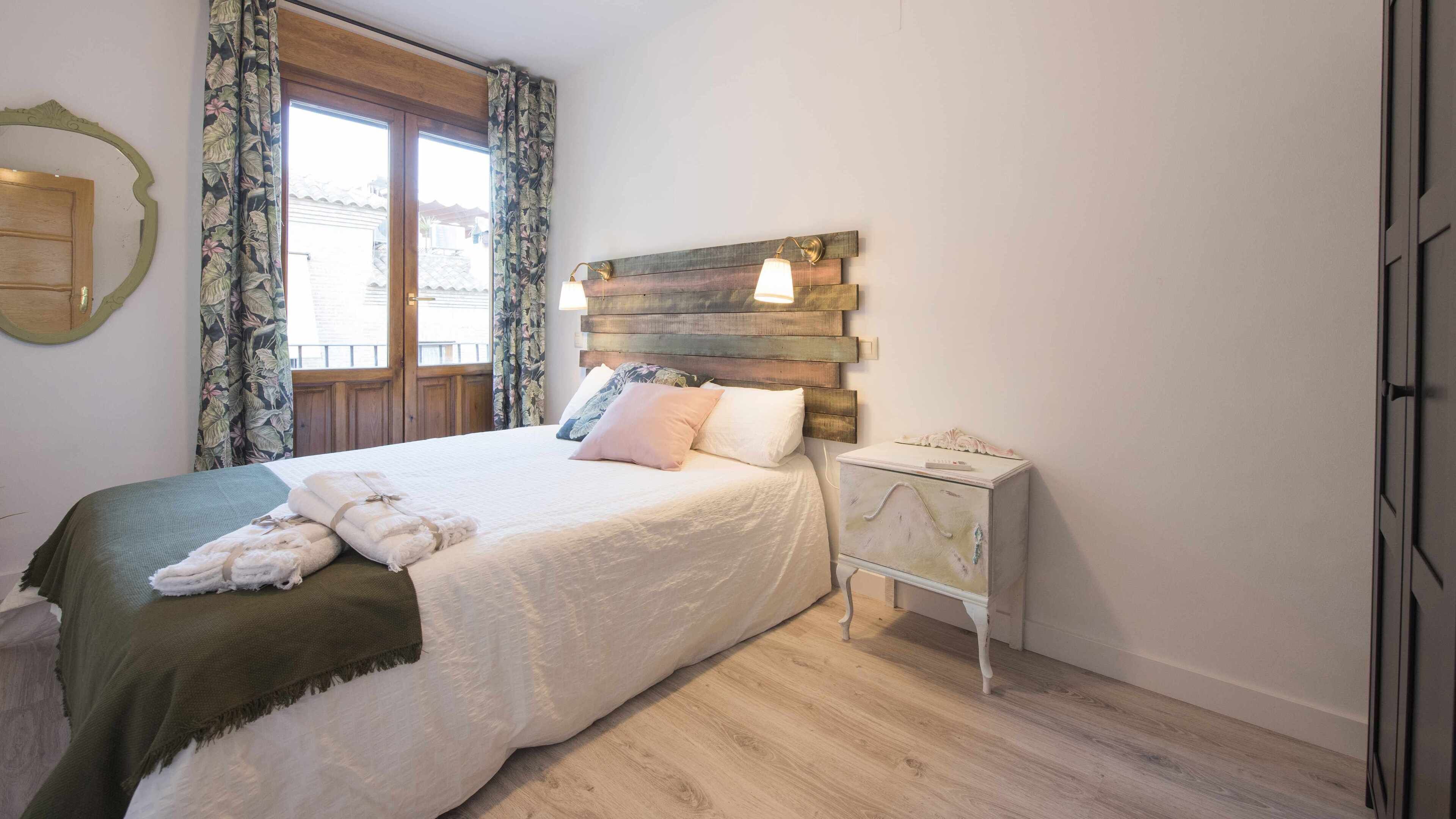 Un dormitorio con cama, mesita de noche, cabecera de madera y ventana con cortinas.