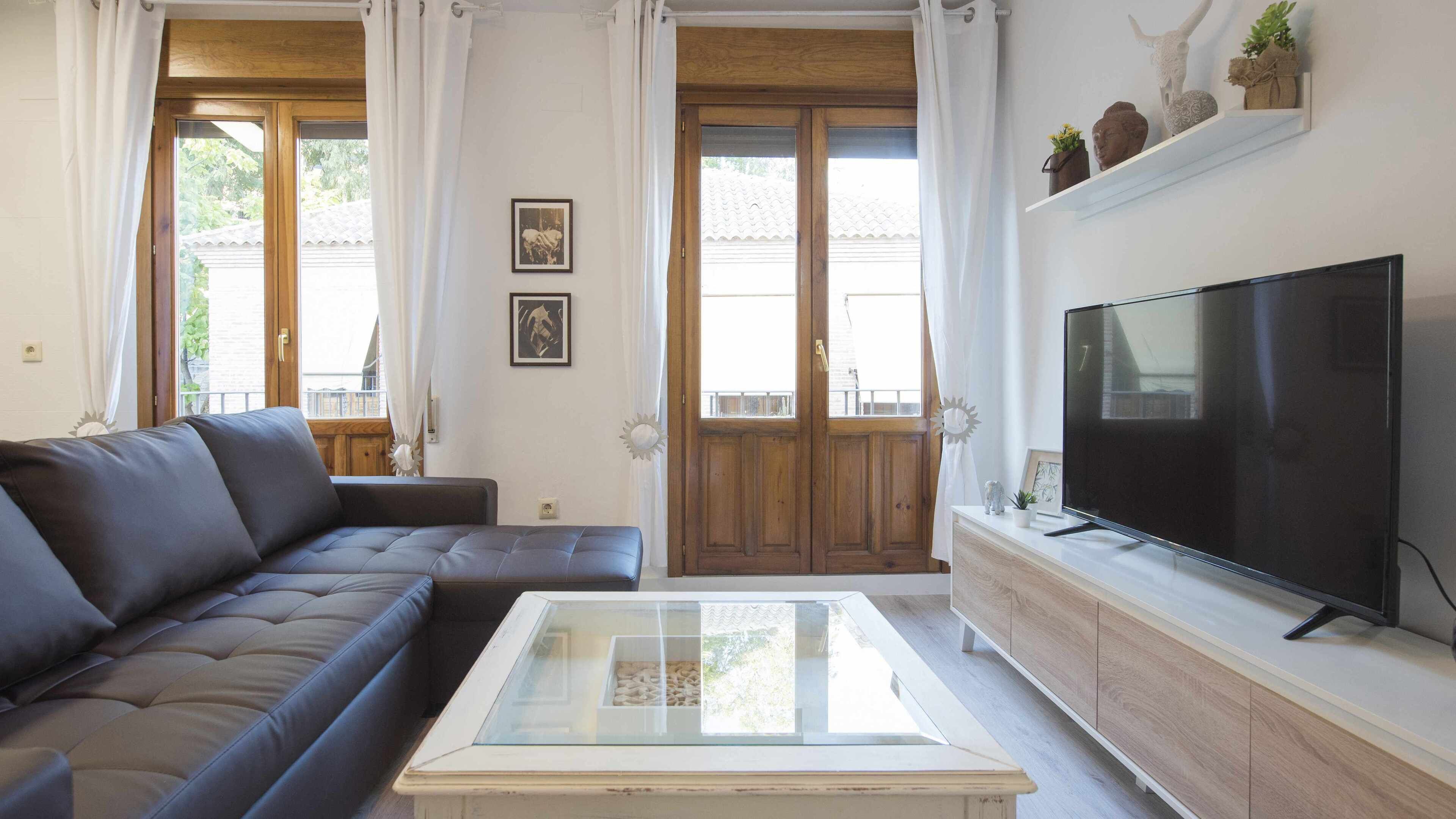 Una sala de estar con un sofá marrón oscuro, una mesa de centro de vidrio, un televisor de pantalla plana sobre un mueble de madera y una puerta de madera con paneles de vidrio.