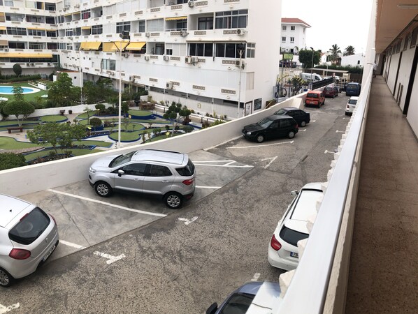 Un estacionamiento con varios autos, un edificio con balcones y un área ajardinada con árboles y una piscina.