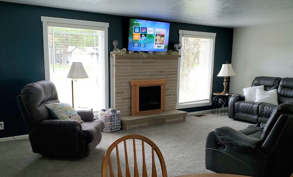 A living room with a fireplace, two leather chairs, a television mounted above the fireplace, and large windows with blinds.