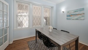 Une salle à manger avec une table et des chaises en bois, de grandes fenêtres équipées de stores, un tapis et un tableau accroché au mur.
