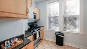 Une cuisine avec des meubles en bois, une cuisinière et un four à micro-ondes. Il y a une fenêtre avec des stores et une poubelle.