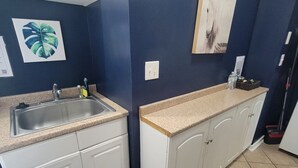 Cocina con gabinetes blancos, encimera de granito, pileta y protector contra salpicaduras. Hay dos interruptores de luz en la pared y un estante con una botella de agua y una planta.