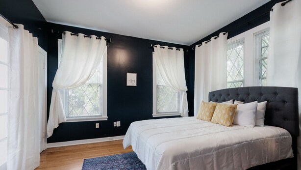 Un dormitorio con una pared oscura, una cama grande, cortinas blancas y una alfombra azul.