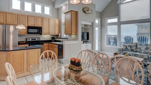 A modern kitchen with wooden cabinets, stainless steel appliances, and a dining area with a glass table and wicker chairs. - Ocean View, steps from sand, enjoy it all in your spacious 3-bedroom retreat (Surf City)