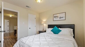Quarto com cama, mesa de cabeceira, abajur, porta e corredor.