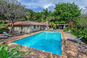 A swimming pool with lounge chairs, a covered seating area, and lush greenery.