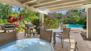 An outdoor patio with a glass table and chairs, a barbecue grill, and a pool in the background.