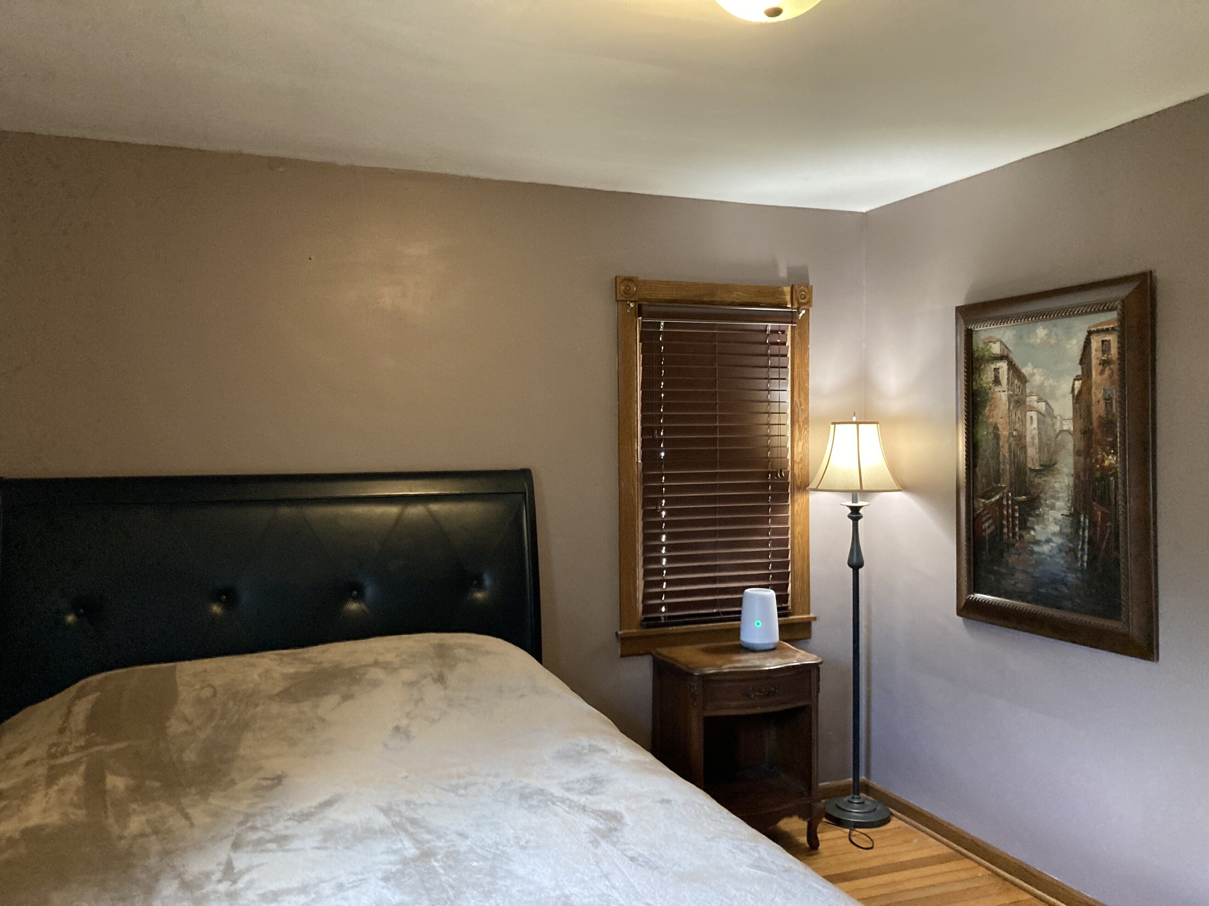 A hotel room with a bed, a nightstand, a lamp, a painting, and a window with blinds.