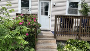 Una casa amb un porxo de fusta, una porta blanca i un petit jardí amb plantes verdes i flors roses.