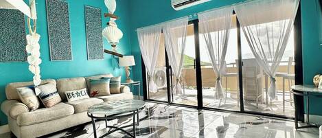 A spacious living room with a teal wall, a marble floor, a sofa with decorative pillows, a coffee table, and a large window with sheer curtains.