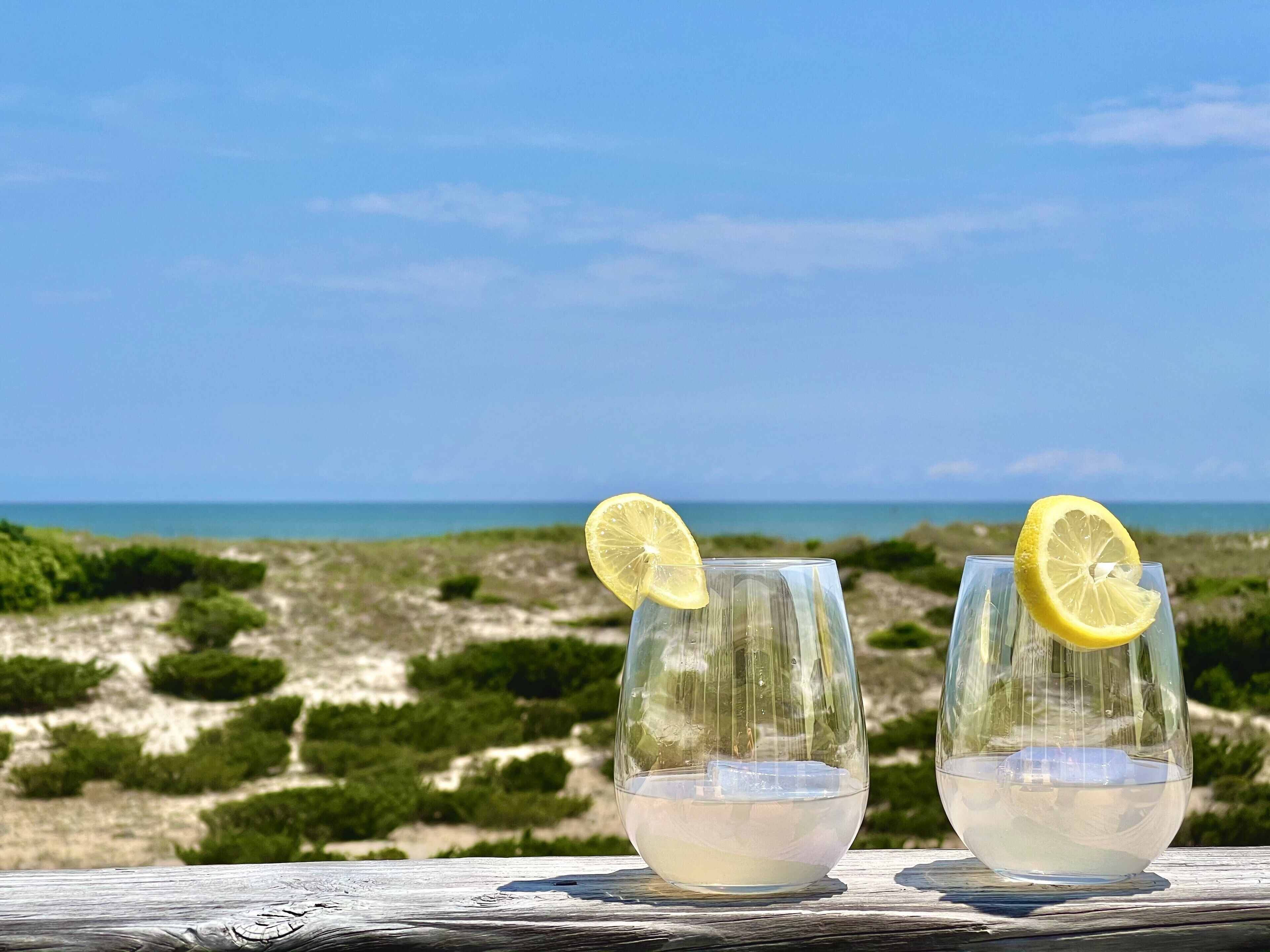 Deux verres d’une boisson claire, décorés de tranches de citron, sont posés sur une surface en bois. On aperçoit une plage et l’océan en arrière-plan.