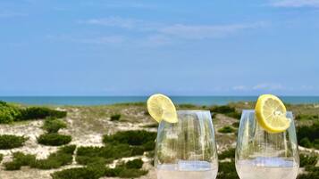Deux verres d’une boisson claire, décorés de tranches de citron, sont posés sur une surface en bois. On aperçoit une plage et l’océan en arrière-plan.
