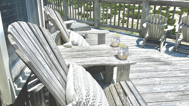 A wooden deck with Adirondack chairs and a table, overlooking a beach with dunes and the ocean.