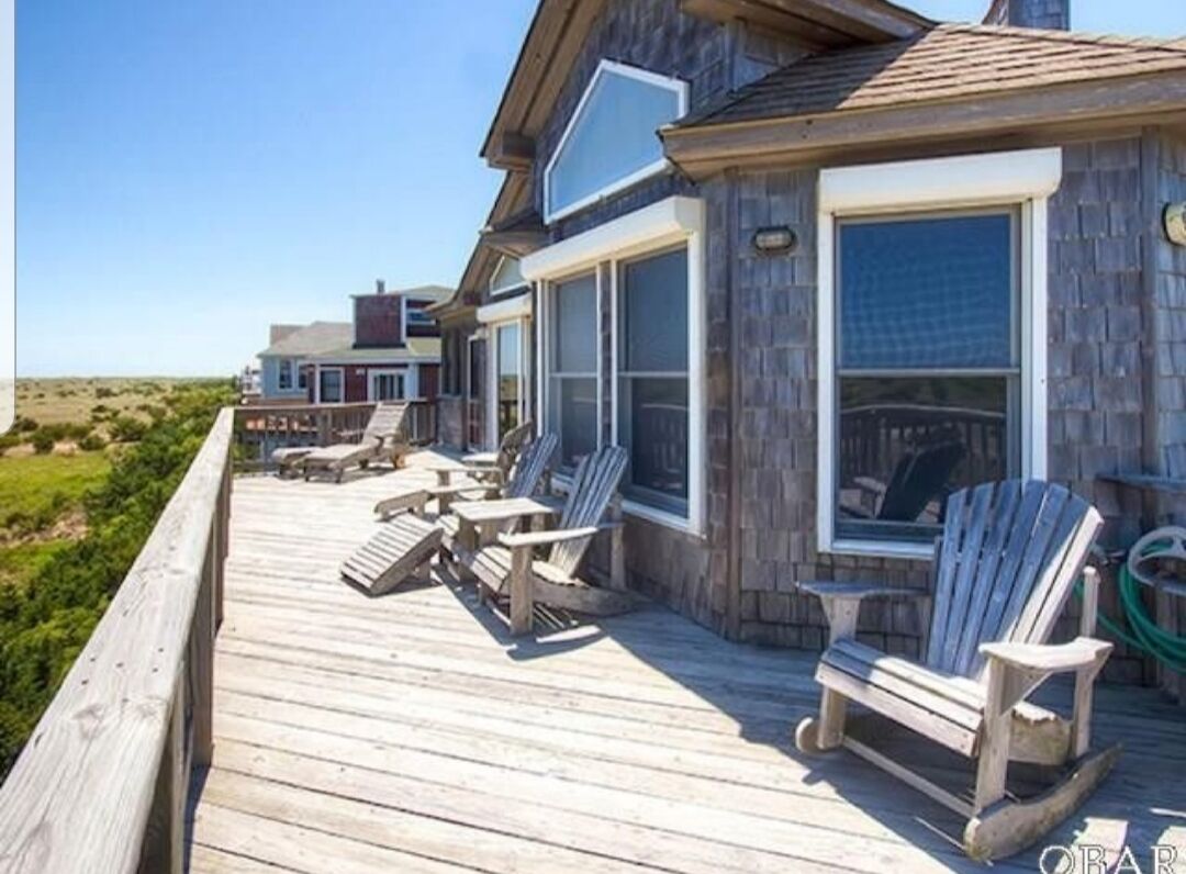 Une terrasse en bois avec des fauteuils Adirondack, une clôture en bois et une maison dotée de grandes fenêtres.