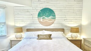 A bedroom with a white brick wall, a round wall hanging of a beach scene, a bed with white bedding, two bedside tables with lamps, and a pillow with "welcome to the BEACH" text.