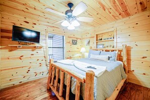 Un dormitorio tipo cabaña de madera con una cama, un televisor montado en la pared, un ventilador de techo y una ventana con persianas.