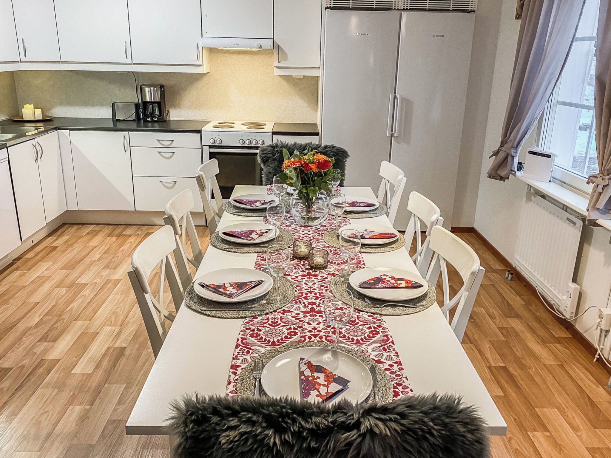 A modern kitchen with white cabinets, a dining table set for six with a patterned table runner, and a fur throw on the floor.