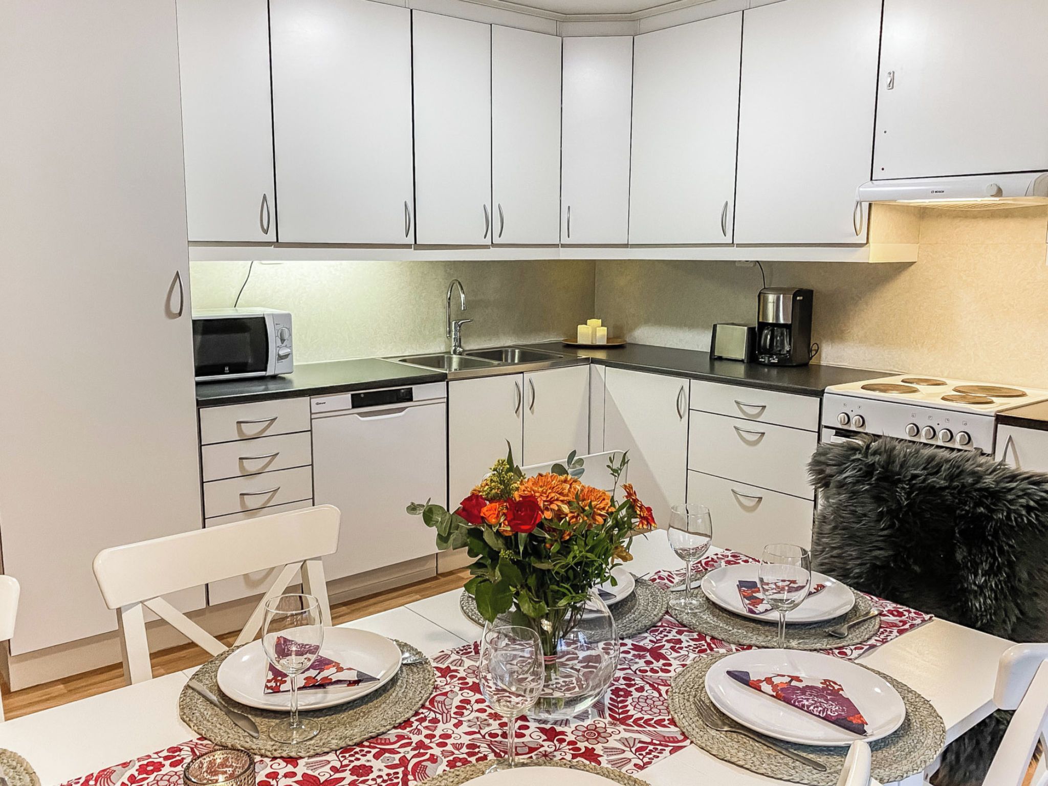 A modern kitchen with white cabinets, a microwave, a coffee maker, and a dining area set for two with a table, chairs, and a vase of flowers.