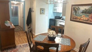 Een eetgedeelte met een ronde houten tafel, stoelen en een schaal eten. Achtergrond is een keuken met een magnetron en kastjes.