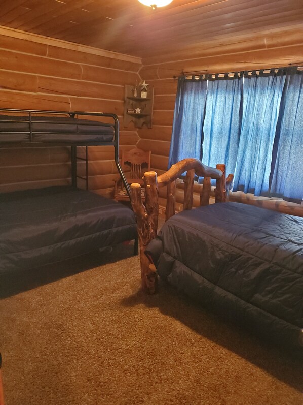 A log cabin room with two bunk beds, a wooden headboard, a small shelf with decorative items, and a window with blue curtains.