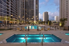 Un espace piscine d’hôtel comprenant plusieurs piscines, des chaises longues et un immeuble de grande hauteur en arrière-plan.
