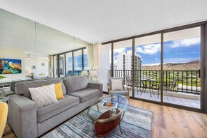 A modern living room with a grey sofa, a glass coffee table, and a balcony with a view of a building and mountains. - 34th floor lanai w/ pool & hot tub access (Honolulu)