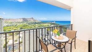 A balcony with a table and chairs, overlooking a coastal landscape with buildings and a mountain.