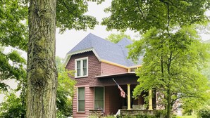 Une maison à deux étages, avec une façade de couleur brun foncé, un toit gris et un porche à l’avant orné d’un drapeau.