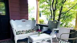 Un ensemble de canapé et de fauteuil en osier, accompagné d’une petite table, est installé sur une terrasse carrelée, offrant une vue sur les arbres et les bâtiments.