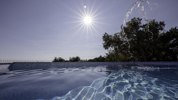 Piscina transparente com reflexos de luz solar na água, rodeada por árvores e um muro de pedra.
