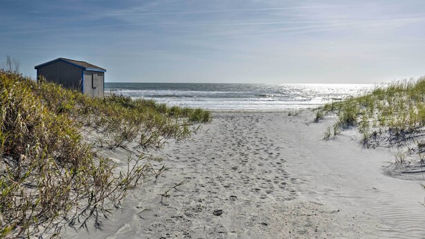 Ein Sandstrand mit einem kleinen Gebäude, vermutlich einer Strandhütte, und Dünen mit Vegetation.