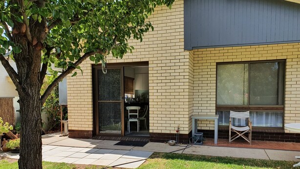 Een woongebouw met een bakstenen gevel, een glazen schuifdeur als inkom, een klein terras met een tafel en stoelen, en een boom op de voorgrond.