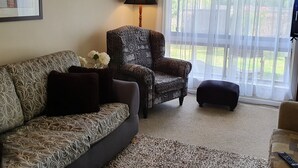 Een woonkamer met een gestreept bankstel, een gestreept fauteuil, een klein poufje, een lamp en een groot raam met dunne gordijnen.