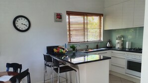 Een keuken met een centraal eiland, een eethoek met stoelen, een klok aan de muur en een raam met zonwering.