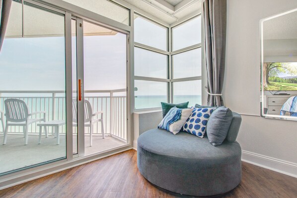 Quarto com uma grande porta de vidro deslizante que dá para uma varanda com cadeiras, um sofá cinza com almofadas estampadas e vista para o mar.