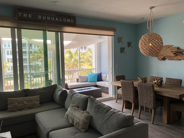 A modern living room with a grey sofa, a wooden dining table, and a balcony with a view of palm trees. - Experience The Bungalow at Margaritaville! (Bradenton)