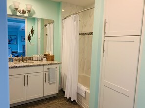 A bathroom with a marble countertop, a white bathtub, a shower curtain, and a mirror with wall art. - Experience The Bungalow at Margaritaville! (Bradenton)