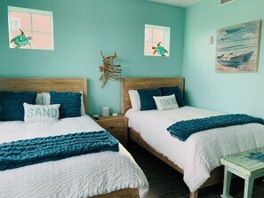 A bedroom with two beds, each with a wooden headboard, white bedding, and teal accents. There are two nightstands with a small table and a decorative piece between the beds. - Experience The Bungalow at Margaritaville! (Bradenton)