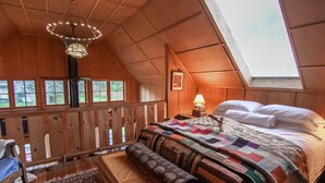 Une chambre mansardée chaleureuse, avec un plafond en pente, une grande fenêtre, un lit recouvert d’une courtepointe, une table de chevet et un banc en bois.